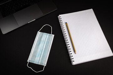 Blank notebook with pencil on black background with laptop computer. Medical mask laying next to notebook. Flat lay image for business concept.