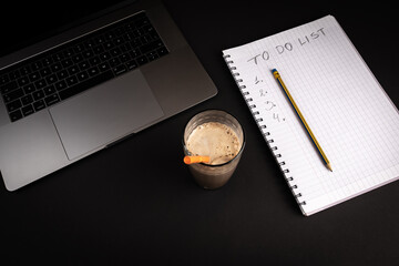 Blank To Do notebook with pencil on black background with laptop computer . Glass of coffee with straw. Flat lay image for business concept.