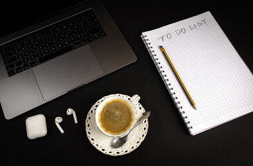 Blank To Do notebook with pencil on black background with laptop computer and bluetooth headphones. Cup of coffee. Flat lay image for business concept.