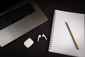 Blank notebook with pencil on black background with laptop computer and bluetooth headphones. Flat lay image for business concept.