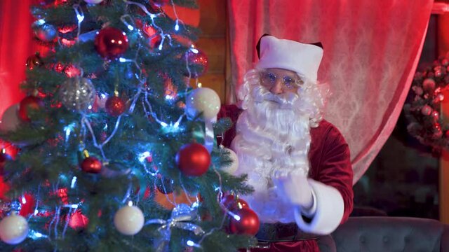Santa Claus near beautiful Christmas tree. Real Santa with white beard admires shiny Christmas balls hanging on a festive tree in New Year.