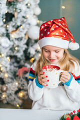 Little smiling girl Santa helper drink hot chocolate with marshmallows from christmas mug with...