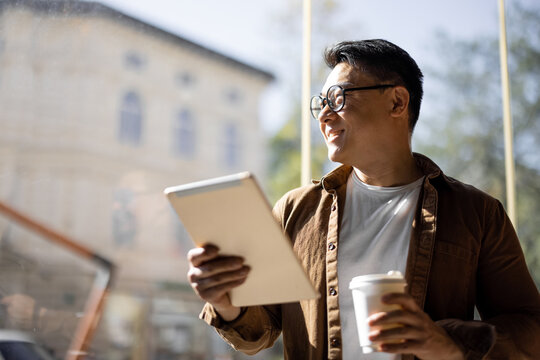 Asian businessman drinking coffee and watching something on digital tablet inside unknown building. Concept of remote and freelance work. Modern adult successful man in glasses. Sunny day