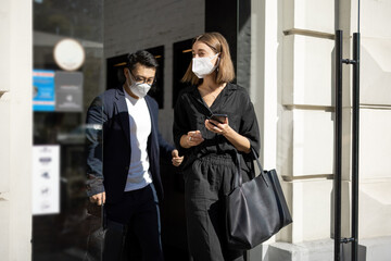Asian man and caucasian woman going from entrance of building. Concept of health protection during Coronavirus pandemic. Smiling modern successful people. Girl with smartphone. City at sunny day