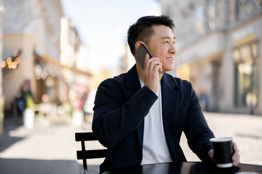 Asian businessman drinking coffee and talking on mobile phone at outdoor cafe. Concept of rest and leisure. Modern adult successful man wearing suit. Blurred background of sunny city street