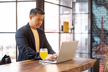 Asian man working on laptop while standing at the bar with a glass of beer. Concept of remote work and business at pub with alcohol. Man wearing formal wear