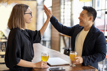 Multiracial friends or business partners having a deal while drinking beer in modern pub. Concept of business cooperation while rest in pub. Pleased young european woman and adult asian man