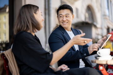 Caucasian woman and asian man having a business conversation while sitting at outdoor cafe. Concept of remote and freelance work while resting. Idea of teamwork and business cooperation