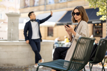 Young woman and asian man using smartphones on city area with fountain. Concept of city life. Beautiful girl wearing glasses sitting on shair. Guy with headphones. Warm sunny day