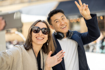 Smiling couple taking selfie on smartphone on city area with fountain. Concept of city life. Idea of friendship. Young beautiful woman and asian man waving hands. Warm sunny day