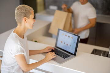 Blonde woman choosing groceries in online store on laptop. Partial of man with shopping bags on background of girl. Concept of online shopping and e-commerce. Idea of healthy eating. Kitchen interior