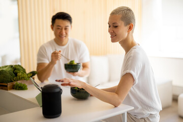 Caucasian woman pushing button on portable smart speaker while eating salad with her husband. Concept of healthy and vegetarian eating and smart home. Smiling multiracial couple in studio apartment