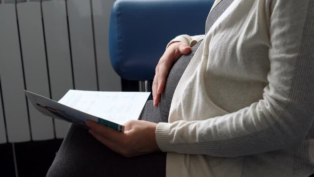 Young Pregnant Woman In Medicine Protective Mask Reading On Couch Waiting For Appointment Medical Control On Hospital. Visit Gynecologist Doctor At Clinic. Examine Expectant Belly Healthcare Check Up