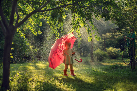 Little cute girl with red umbrella under beautifully lighted summer rain in the park. Image with selective focus and toning