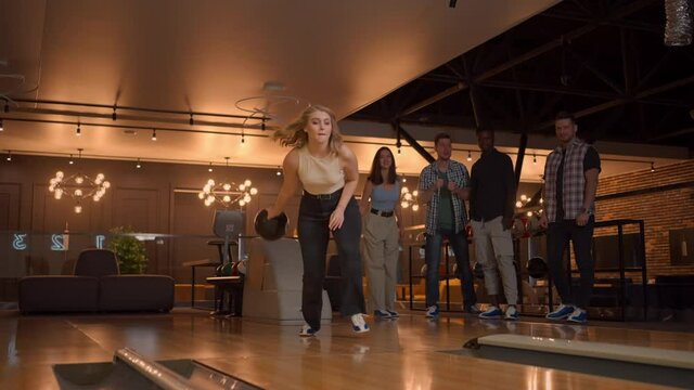 A young woman in bowling throws a ball on the track and knocks out a shot in slow motion and jumps and dances for joy. Friends fans support and clap with a smile.