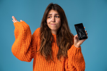 Portrait of a pretty sad woman in orange jumper holding smartphone with broken screen and throwing...