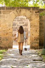 Fototapeta na wymiar mujer preciosa andando de espaldas por un camino de piedras y cesped con un bolso, chaqueta marron y camisa blanca en Mallorca