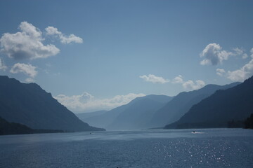 Teletskoye Lake in the Altai Mountains. The water glistens in the sun. Trees grow on the mountain. Blue sky and white clouds