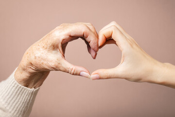 Hands of a young and senior woman make a heart gesture. Close up. Beige background. Concept of care