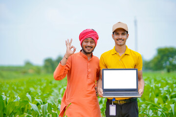 Young indian banker or agronomist showing laptop screen with farmer at agriculture field.