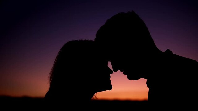 Closeup silhouettes of man and woman in love looking at each other. They are standing in front of violet and yellow sky touching their foreheads. then the image blurs.