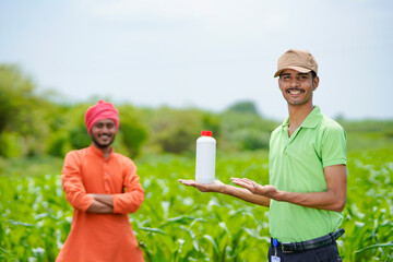 Young indian agronomist holding liquid fertilizer bottle with farmer at green agriculture field.
