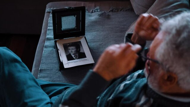 lonely old man sits on a sofa at home and opens a box with his old pictures from the past