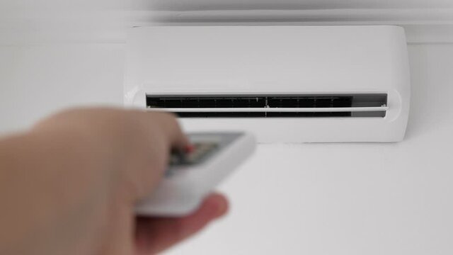 Flat air conditioner on white wall in room interior. Man pushing button on remote. Setting temperature