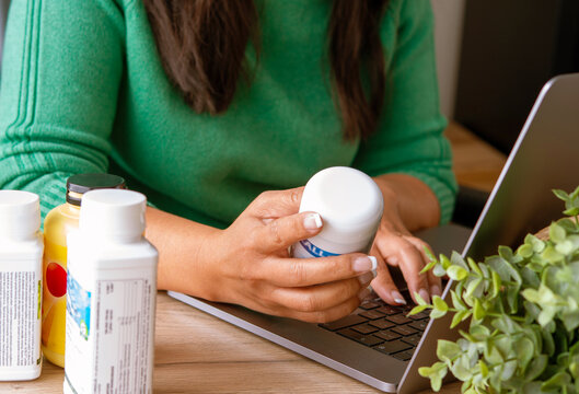 Health and nutrition concept. Indoor image young female holding  food supplement bottle sitting with laptop surfing searching internet for online review of food supplements products on internet.
