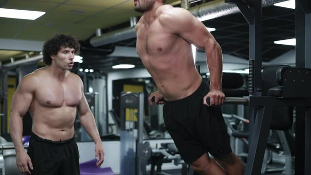 Trainer Coaches Client in Gym. Young Athletic Man doing push-ups on parallel bars.