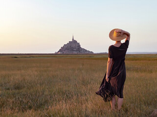 Young woman wearing hat a dress running towards mont saint michel  - obrazy, fototapety, plakaty
