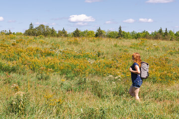 Caucasian, red headed adult woman in forties walking in summer field against blue sky. Has backpack and shorts on. Pale legs. Exploration. 