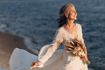 Fototapeta na wymiar Happy senior woman in wedding dress. Portrait of senior bride with bouquet on seashore.