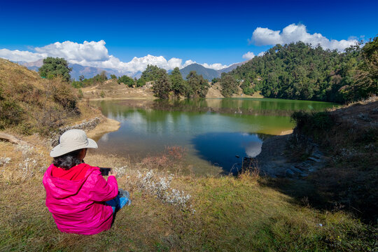 Solo traveller, single female tourist taking picture of Deoria Tal with her mobile phone, high altitude lake in Uttarakhand, India. Blue sky with snow-covered mountains in background.