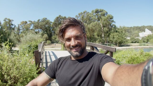 POV of smiling man taking selfie while having break from workout. Front view of happy bearded athlete taking photo in background of beautiful view of summer park. Emotion, sport concept