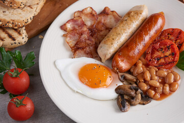 Traditional full English breakfast with fried eggs, sausages, beans, mushrooms, grilled tomatoes and bacon on a plate, Toast, butter, jam on a wooden board