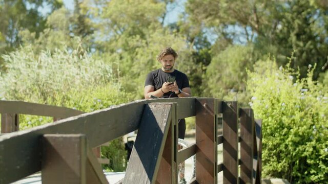 Long shot of man standing on bridge with phone during cycling. Serious Caucasian biker in sportswear using navigator and map app on smartphone, inspecting area. Technology, sport concept