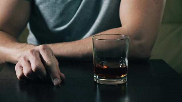 Drunk man wearing casual grey t-shirt looks at wedding ring after quarrel hesitating and decides to divorce drinking glass of whiskey closeup.