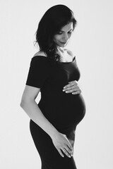 Beautiful young pregnant woman embracing belly posing in studio, black and white photo. Happy...