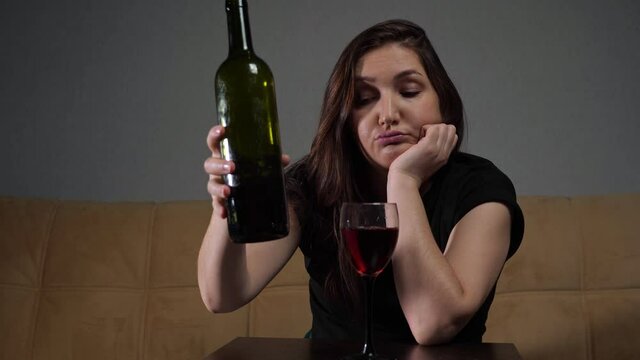 Young brunette drunk woman with depressed face expression pours wine from bottle into wineglass sitting on soft sofa cushion in comfortable living room.