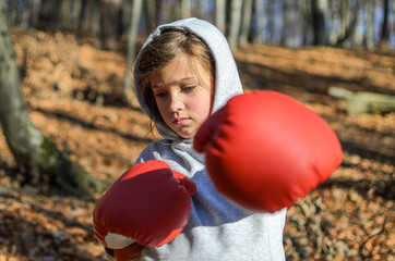 Little adorable girl child in a tracksuit, with a hood on his head, in red boxing gloves, boxing on the street in the autumn forest, young boxer