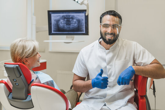 Successful confident Middle-Eastern male dentist orthodontist stomatologist looking at camera showing thumb up while patient client waiting for tooth teeth filling cleaning