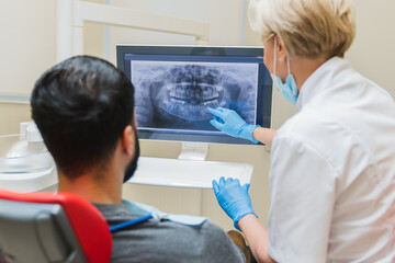Female dentist orthodontist showing explaining an x-ray photo of jaws on computer screen to a male patient in dental clinic. Stomatology and healthcare concept