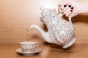 Juego de té con adornos dorados. Mano sosteniendo una tetera vertiendo agua caliente en una pequeña taza de té. Fondo de madera