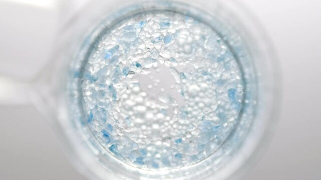 Crystals of blue salt fall down from test tube into clear liquid in flask creating different sized bursting bubbles on light grey background | Mineral skin care cosmetics formulation concept