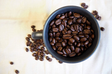 on the beige background of the table with scattered coffee beans, there is a dark mug filled with roasted whole coffee beans. top view