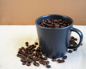 on the beige background of the table with scattered coffee beans, there is a dark mug filled with roasted whole coffee beans. side view. energy drink