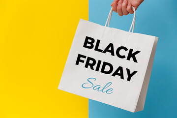 Female hand holding white shopping paper bag with Black Friday Sale text on yellow and blue background. Sale, discount, shopping concept.