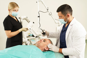 Doctor dermatologist with his assistant working on middle aged male client's laser skin resurfacing...