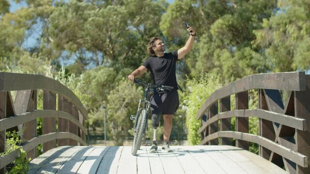 Long shot of biker with disability taking selfie in summer park. Front view of happy man with bionic leg standing with bicycle and taking photo on phone for social network. Disability, sport concept